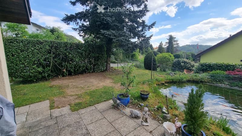 Garden with a pond, small statues, potted plants, and a grassy area surrounded by trees and hedges.