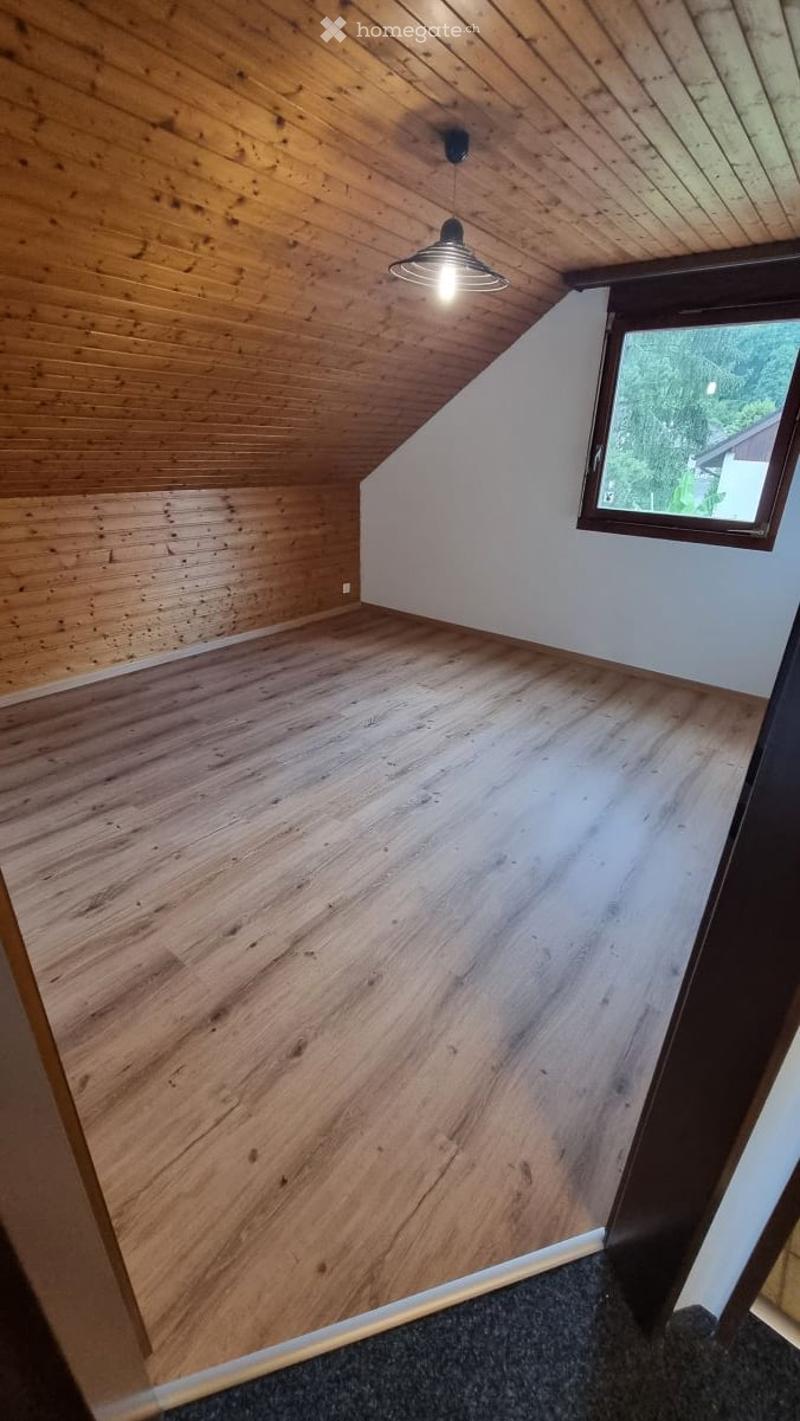 Empty attic room with wooden walls and floor, exposed wooden ceiling beams, and a single window letting in natural light.