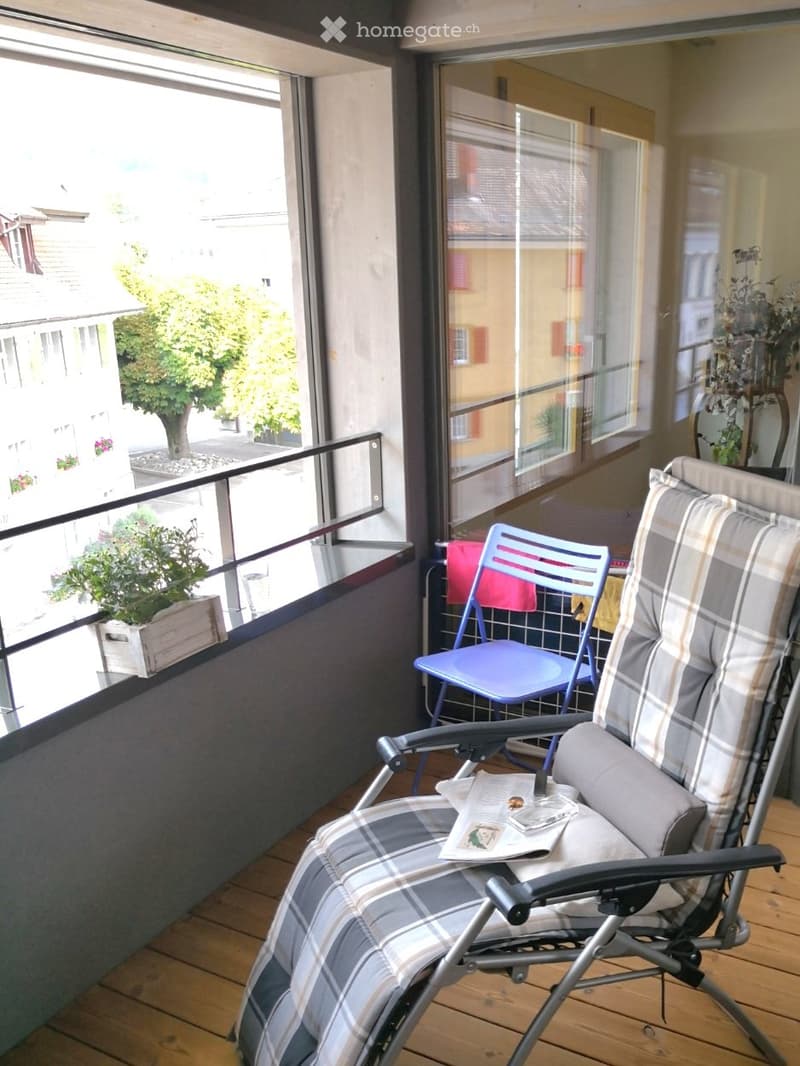 wooden floor, lounge chair, small table, blue chair, railing, potted plants, distant buildings, tree