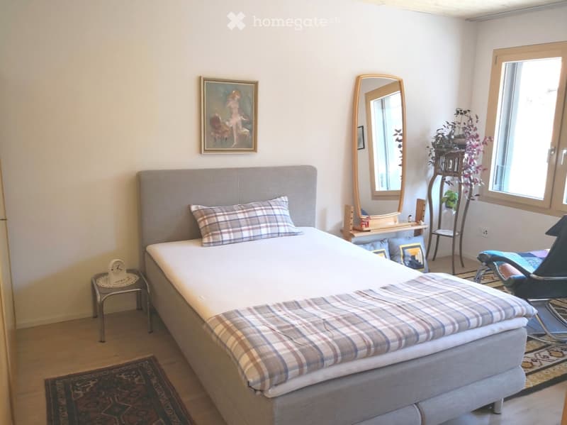 Single bed with grey headboard, plaid pillow, and bedspread, mirror, bedside table with a clock, framed artwork, wooden floor, and a rug.