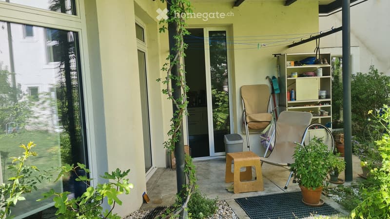 Balcony with a small seating area, including a chair, stool, and potted plants. A shelf with various items is visible, and a clothesline is attached to the wall.