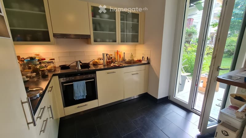 Modern kitchen with light-colored cabinets, stainless steel appliances, and a black tiled floor. The kitchen features an oven, sink, and various kitchen items on the countertops.