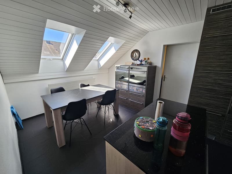 A modern, well-lit room with a dining table, chairs, and a built-in oven. The room features dark flooring, white walls, and skylights providing natural light.