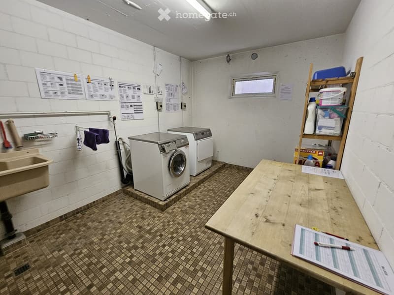 Utility room with white brick walls, tiled floor, washing machine, dryer, wooden table, shelf, and sink.