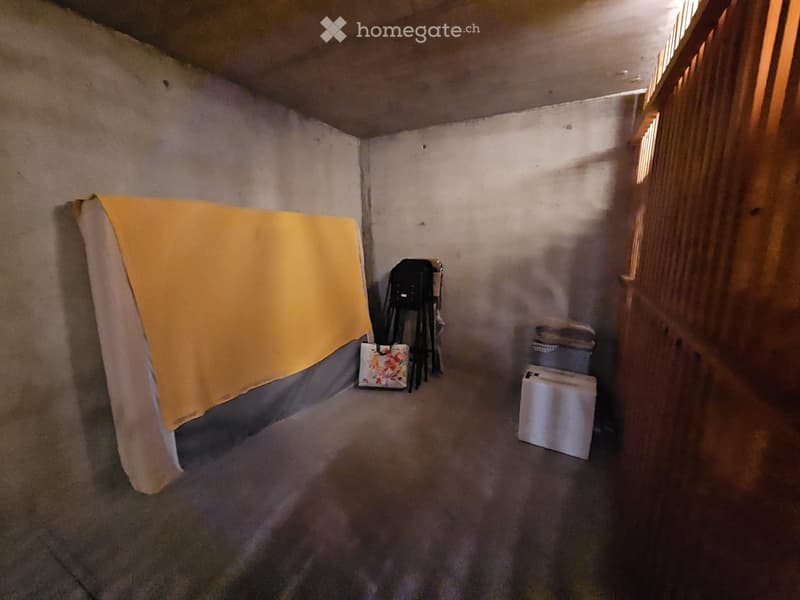 A small, dimly lit room with a bed covered in a yellow blanket, a chair, and a handbag placed against the wall.