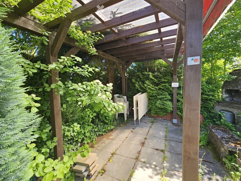 A wooden pergola with a partially open roof, surrounded by lush greenery and plants. There are a few chairs and a table under the pergola, and a stone pathway leads through the garden.