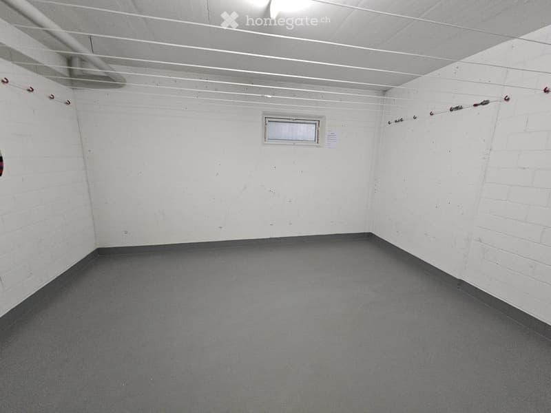 Empty room with white walls, grey carpeted floor, and a small window with a vent. Exposed ceiling with visible pipes and lighting fixtures.
