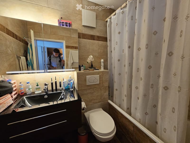 Modern bathroom with a black sink, chrome faucet, white toilet, and a large mirror above the sink. The shower area is enclosed with a white curtain featuring a geometric pattern. Various toiletries and a small flower arrangement are placed on the sink cou
