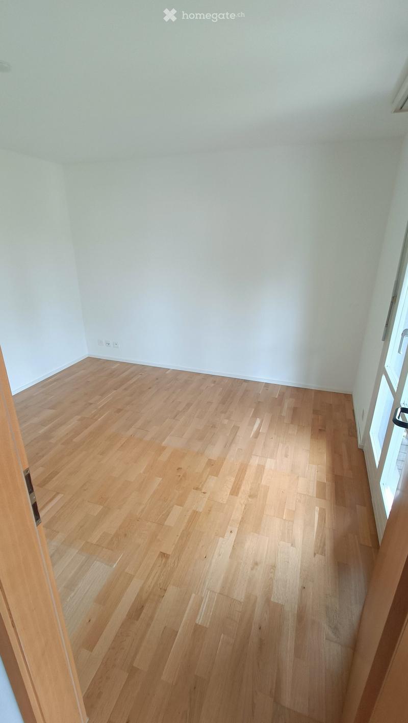 Empty room with light wooden flooring and white walls, featuring a doorway on the left and a window on the right.