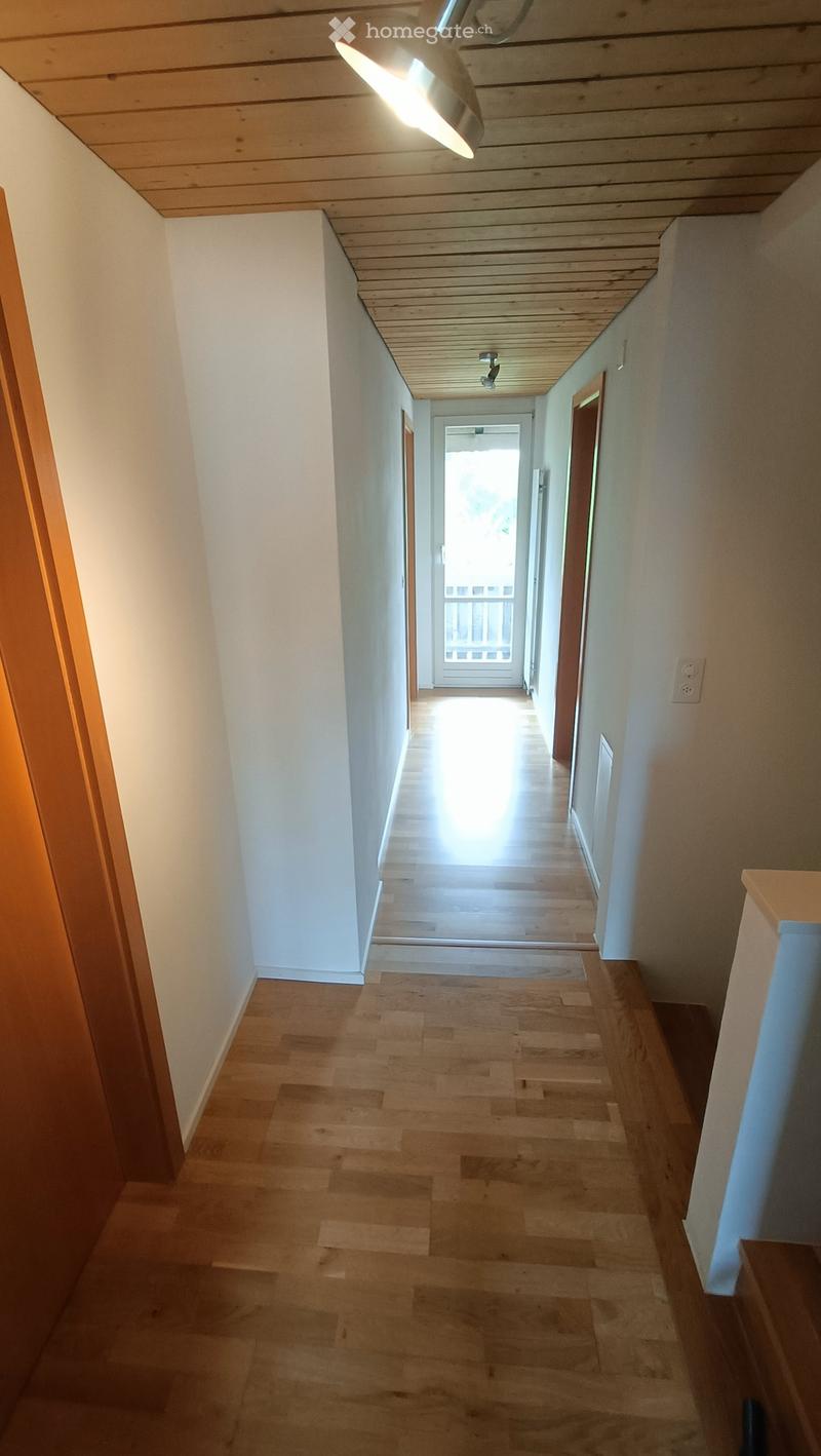 A long, narrow hallway with wooden flooring and white walls, leading to a bright open area at the end.
