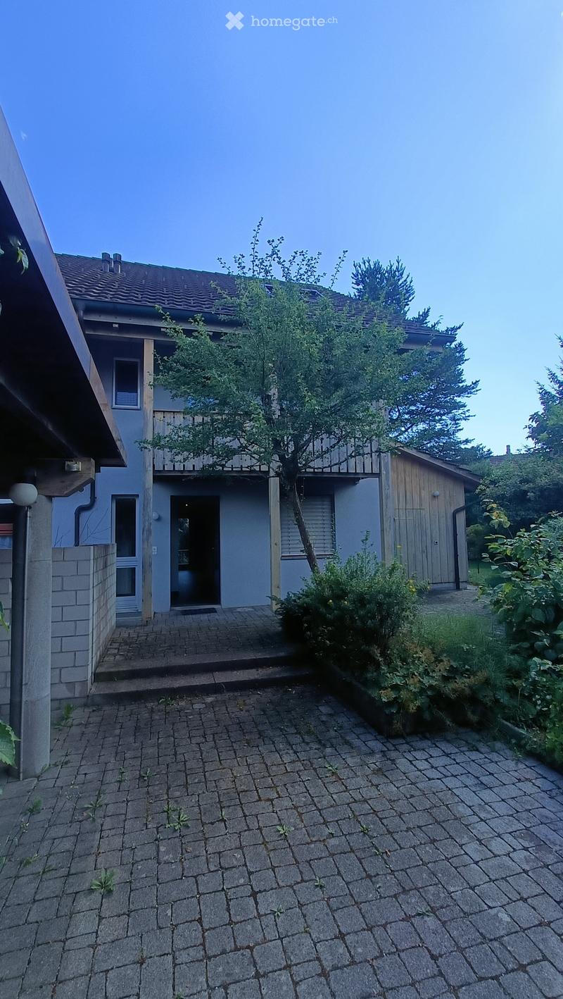 Brick-paved outdoor area with a small tree, bushes, and a two-story house with a balcony and garage in the background.