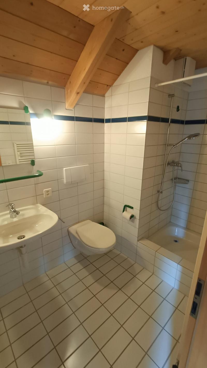 Cozy bathroom with white tiles, a sink, toilet, and a walk-in shower with a glass door. The room features a slanted wooden ceiling and a small window with a blue accent strip.