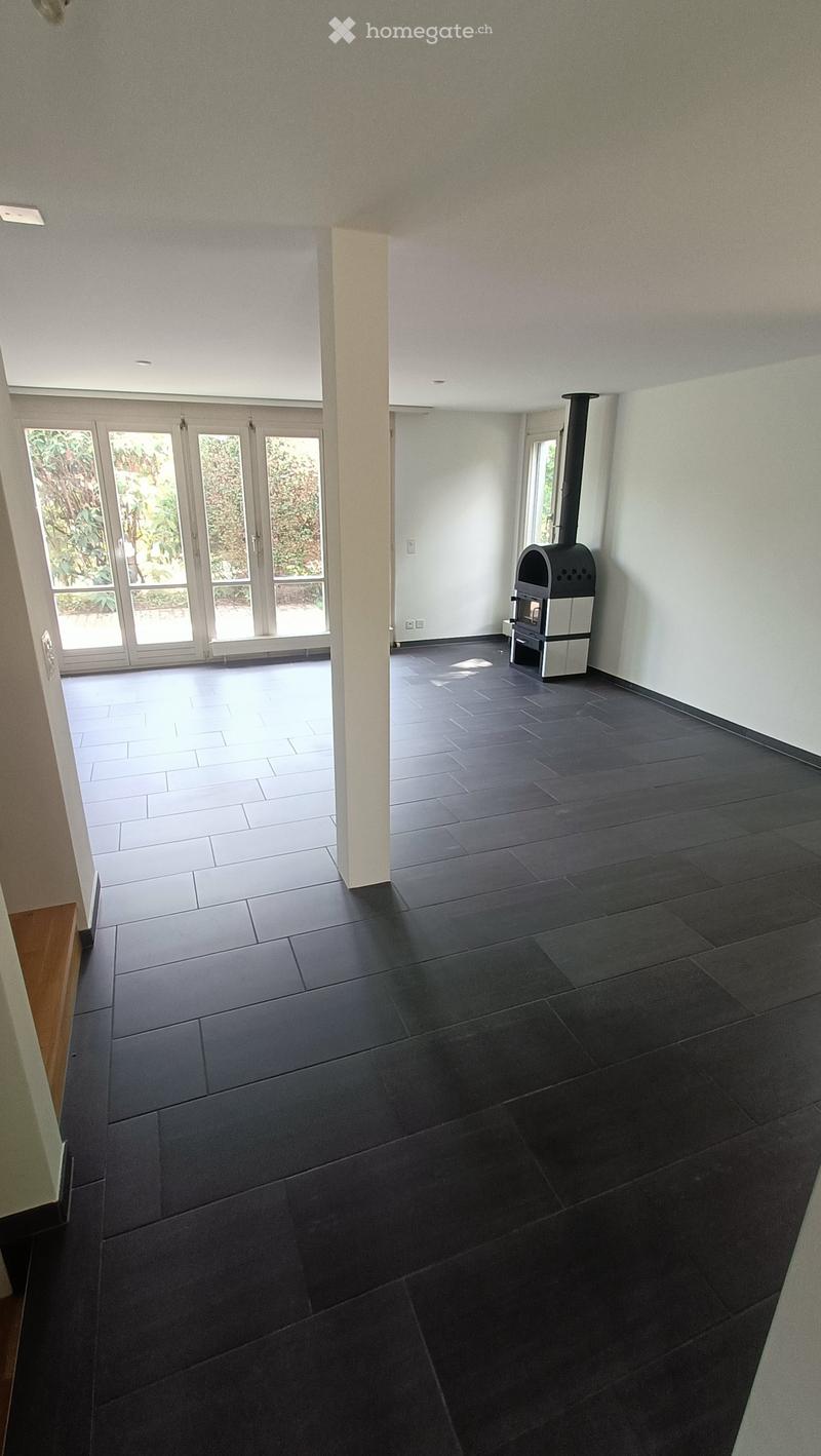 An unfurnished room with dark tiled flooring, white walls, and a modern wood-burning stove. The room features large glass doors leading to an outdoor area and a raised ceiling with recessed lighting.