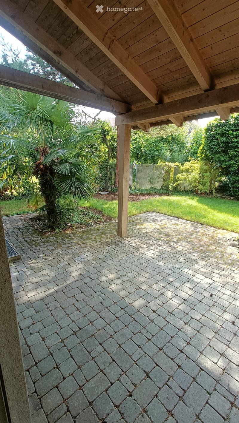 A covered outdoor area with a stone-paved floor, wooden beams, and a lush garden with palm trees and greenery.