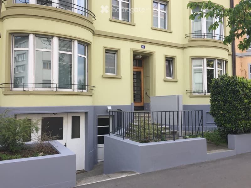 Two-story residential building with light yellow facade, multiple windows, and a raised ground floor entrance with a black railing and small garden area.