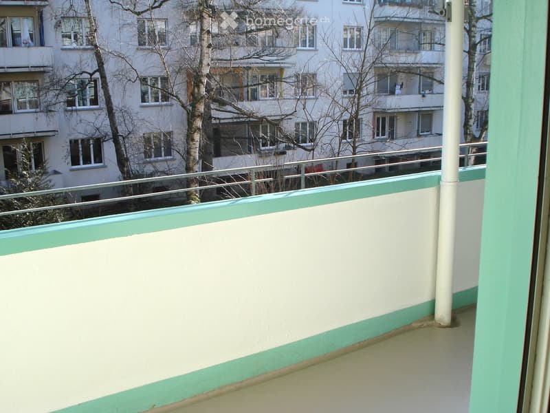 Empty balcony with white walls, green trim, metal railing, and view of apartment building and trees.