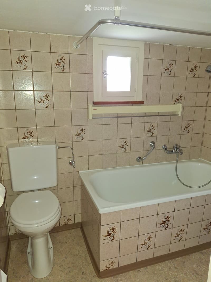 Bathroom with beige tiled walls, a toilet, a bathtub, a shower handle, and a handrail for accessibility.