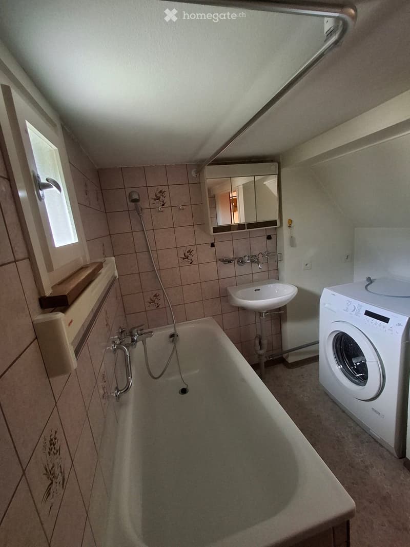 Bathroom with a bathtub, sink, mirror, and washing machine. The room has tiled walls and a window providing natural light.