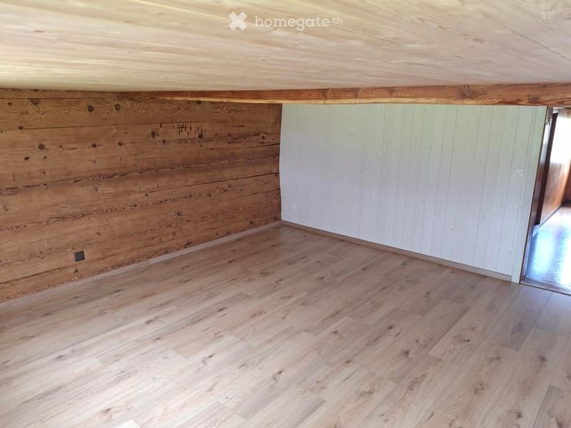 Empty room with wooden flooring, exposed wooden beams on the ceiling, and a mix of light and dark wood paneling on the walls.