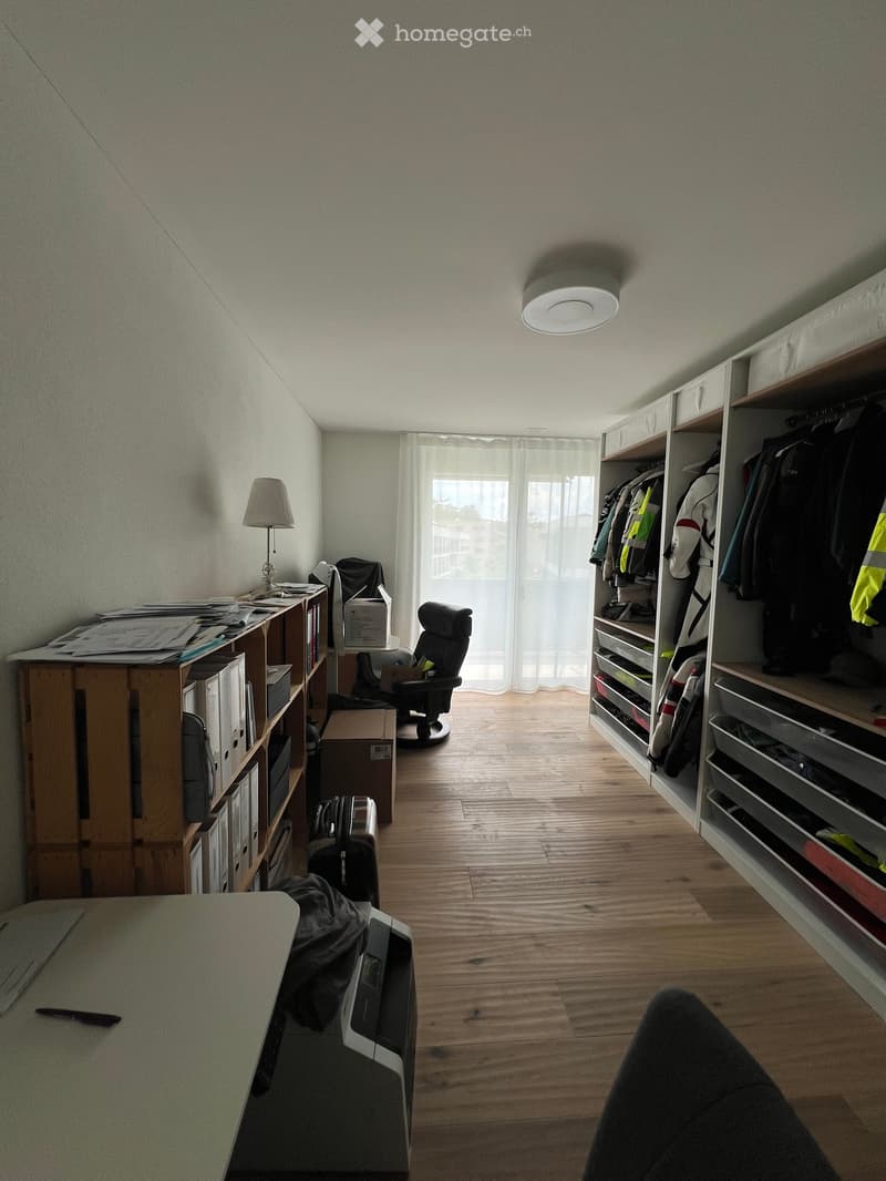 A furnished room with wooden flooring, a large window with white curtains, a chair, a table, a lamp, shelves, and a suitcase.