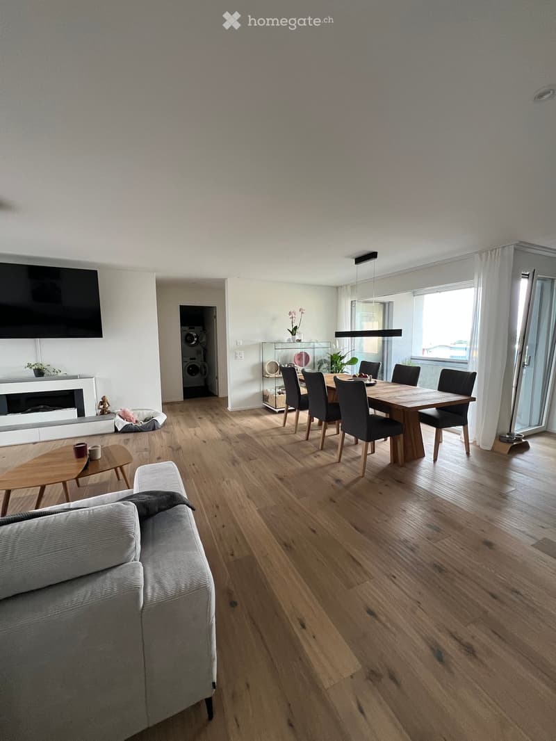 Open-plan living and dining area with wooden flooring, a grey sofa, coffee table, dining table with chairs, fireplace, and a washing machine in the background.