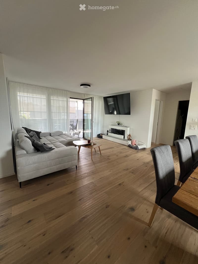 Modern living room with hardwood floors, large windows, a grey sectional sofa, a coffee table, a fireplace, and a TV mounted on the wall. A dining area with chairs and a table is visible.