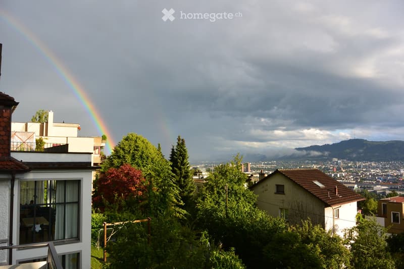 View from a balcony showing a cityscape with trees, houses, and a rainbow in the sky.
