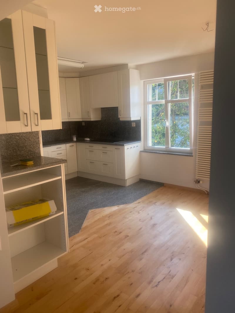Empty kitchen with white cabinets, granite countertops, wooden flooring, and a window with a view of greenery.