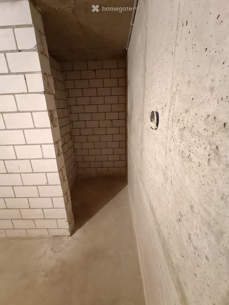 Empty room with bare walls and concrete floor, featuring a small window and a light fixture on the wall.