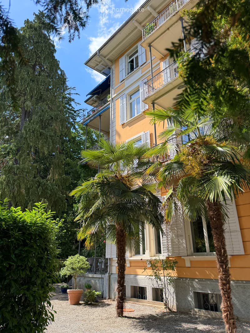 A multi-story yellow building with white shutters and balconies, surrounded by lush greenery and palm trees.