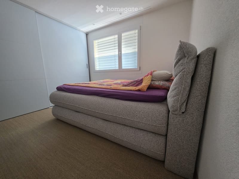A modern bedroom with a grey upholstered bed, colorful blankets, and pillows, set against light-colored walls and carpeted flooring.