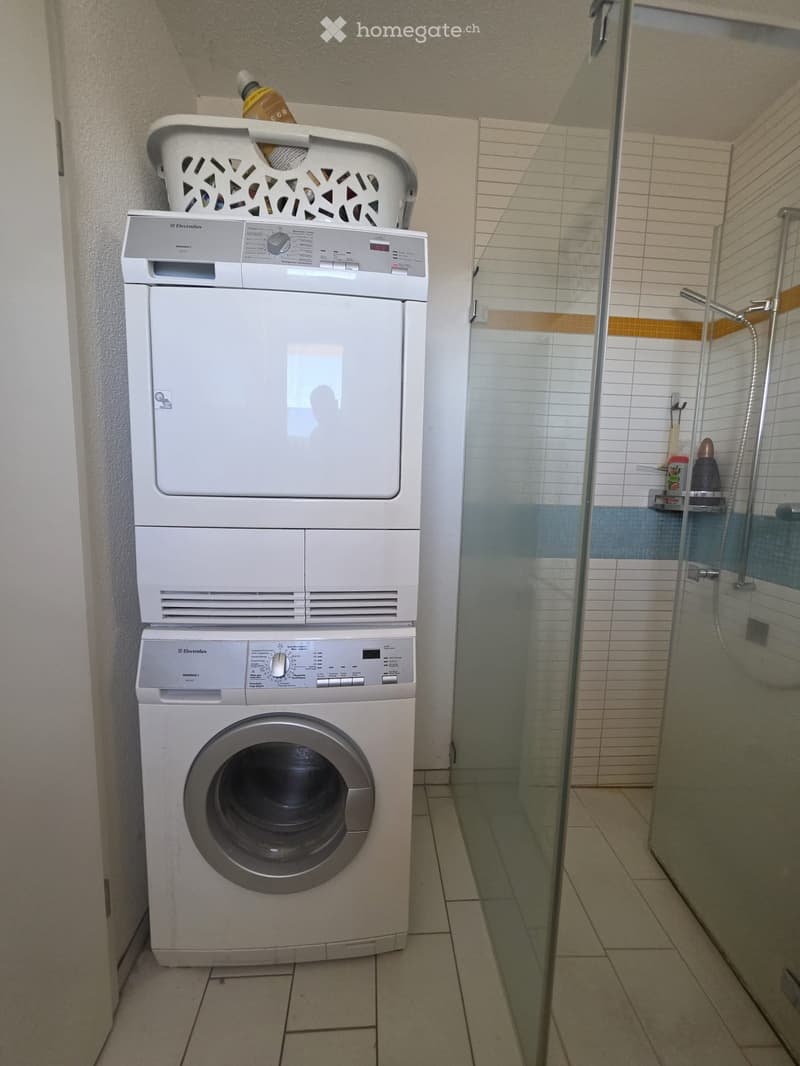 Bathroom with a stacked washing machine and dryer unit, a glass-enclosed shower with a shelf, and tiled walls.