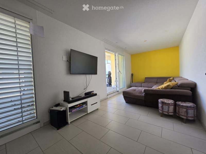 Modern living room with a grey sectional sofa, ottoman, two wooden stools, flat-screen TV mounted on the wall, and a white TV stand with a speaker.