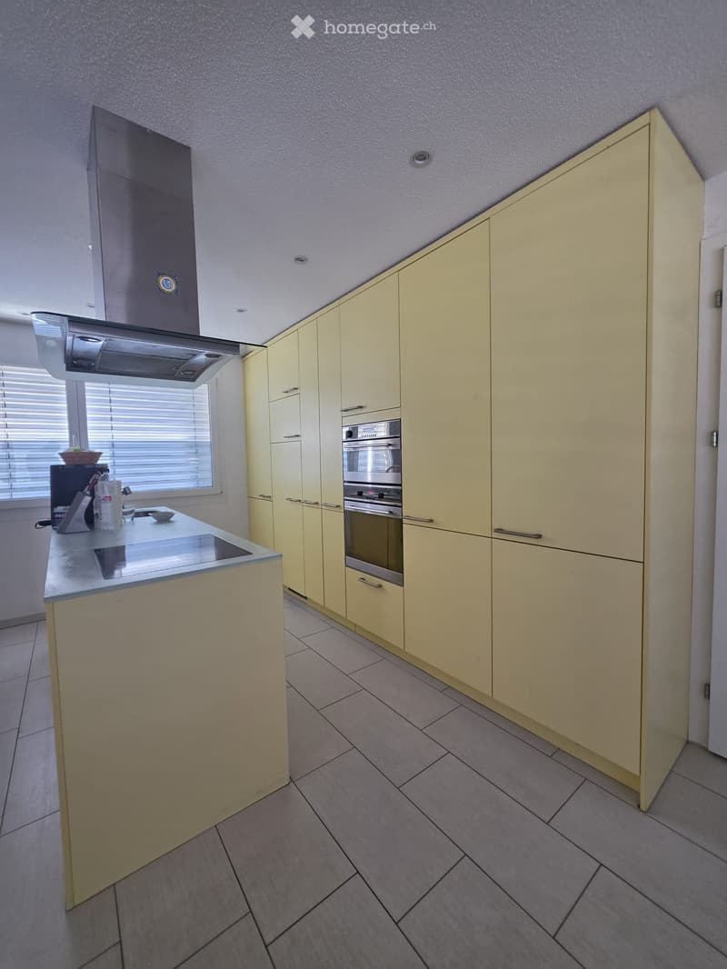 Modern kitchen with light yellow cabinets, stainless steel oven, island with sink, and various small appliances on the counter.