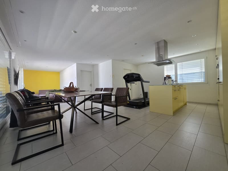 Open-plan room with a dining table, chairs, a kitchen counter, and a treadmill. The space features tiled flooring, large windows, and a yellow accent wall.