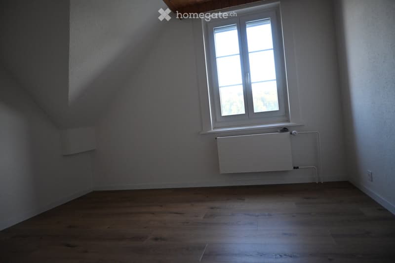 Empty room with wooden flooring, white walls, a window with white frame, and a radiator below the window.