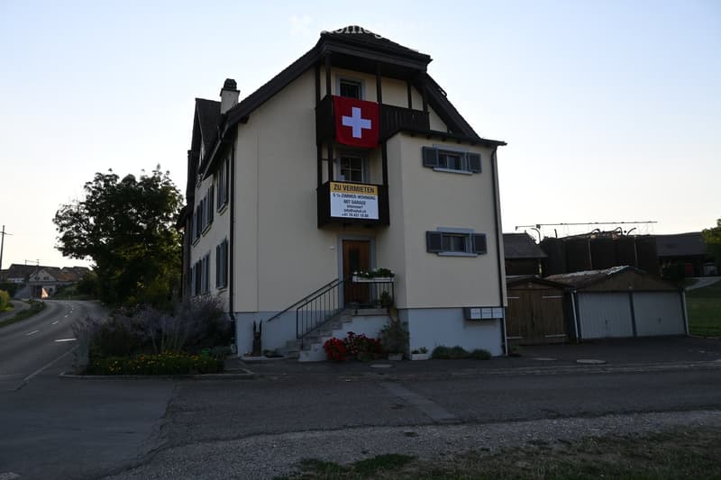 Two-story house with beige siding, multiple windows, a small balcony with a Swiss flag, and a sign advertising a5-room apartment with garage.