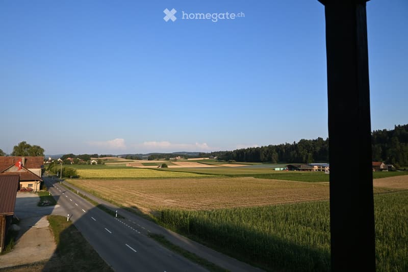 View from a window showing a rural landscape with fields, a road, and scattered houses under a clear blue sky.