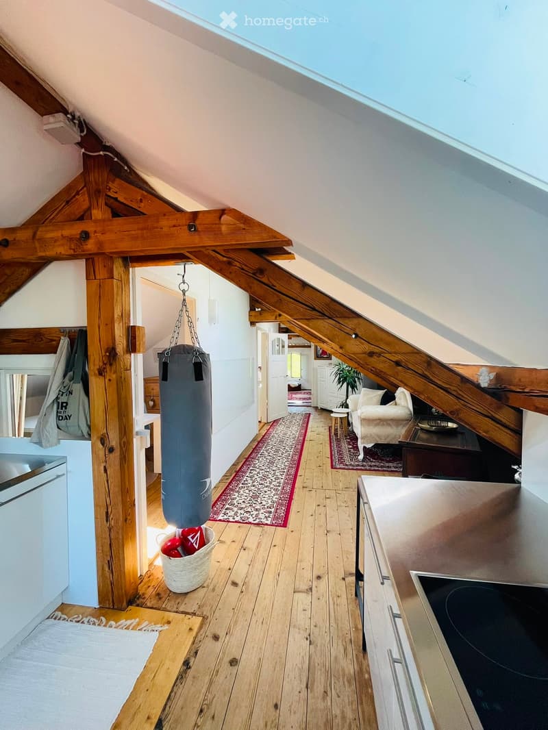 Loft-style room with wooden beams, exposed wooden floor, kitchen area, living area with sofa and small table, punching bag hanging from the ceiling, and decorative rugs.