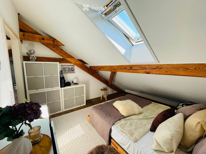 Cozy attic bedroom with wooden beams, skylight windows, a bed with pillows and blankets, a nightstand, a small stool, a cabinet, and a vase with flowers on a table.