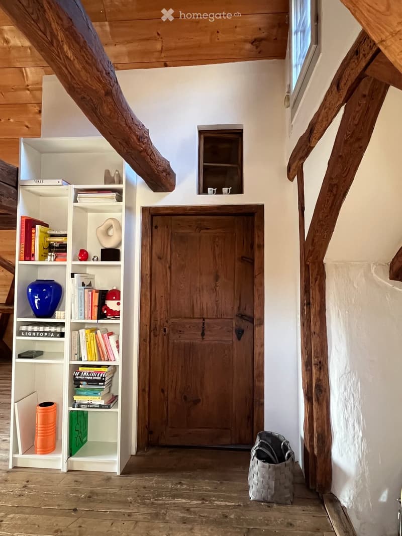 A rustic hallway with wooden beams, a wooden door, a white bookshelf filled with books and decorative items, and a woven basket on the floor.