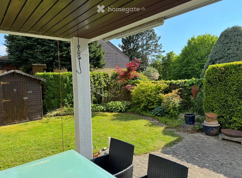 Well-maintained garden with a covered patio area, green lawn, various shrubs and trees, and outdoor furniture including a table and chairs.