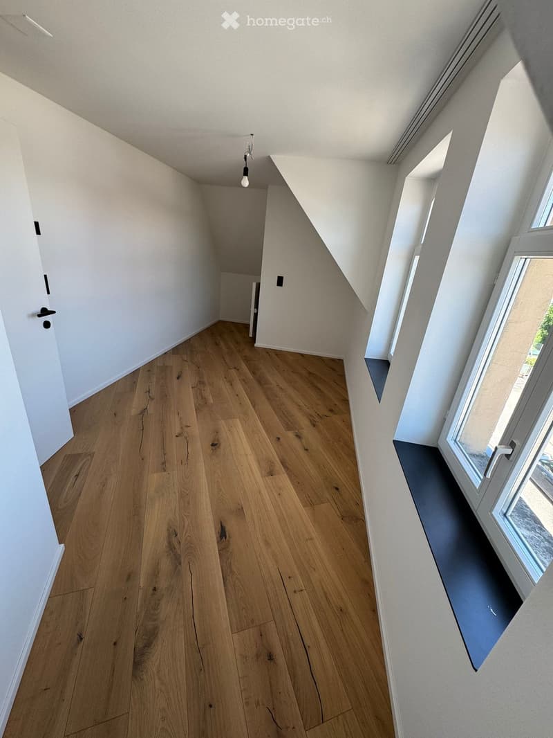 Empty room with wooden flooring, white walls, and a large window on the right side. The ceiling has a sloped design typical of an attic space.