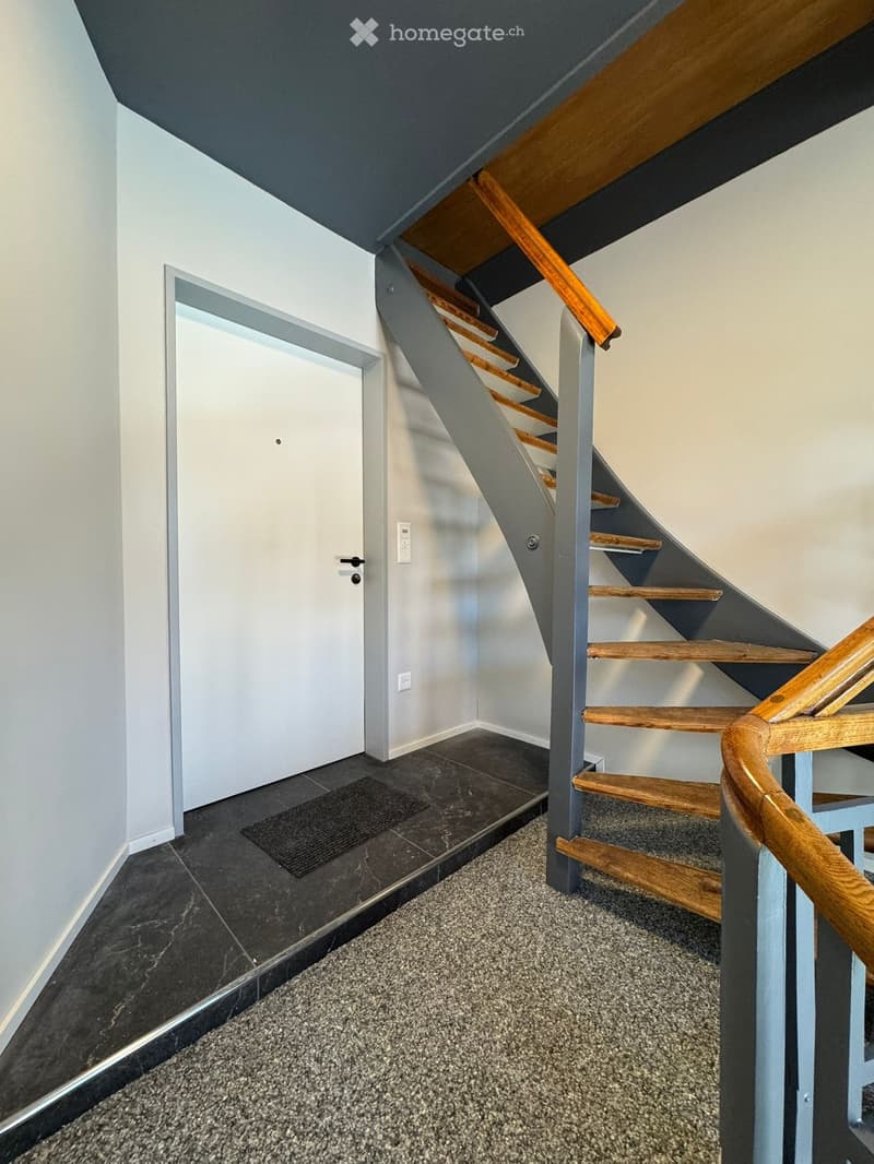Modern hallway with a wooden staircase, grey walls, and a mix of dark tiled and carpeted flooring.