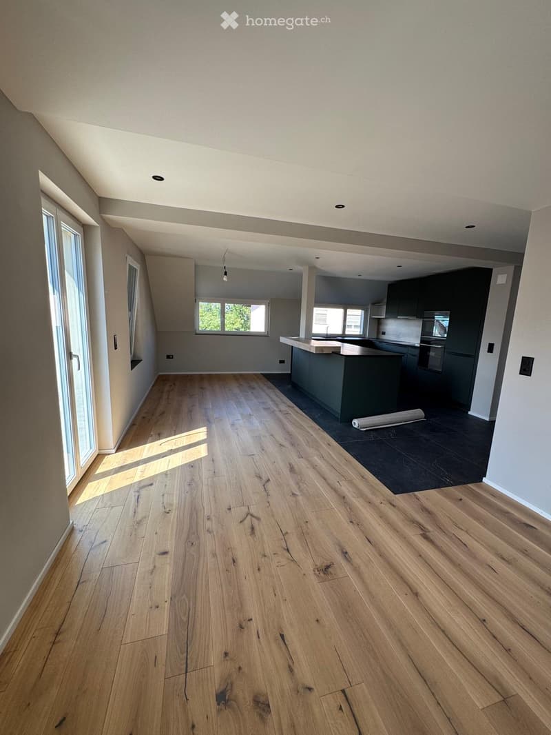 Empty room with light wooden flooring, large windows, and a view of the kitchen area with dark cabinets and a countertop.