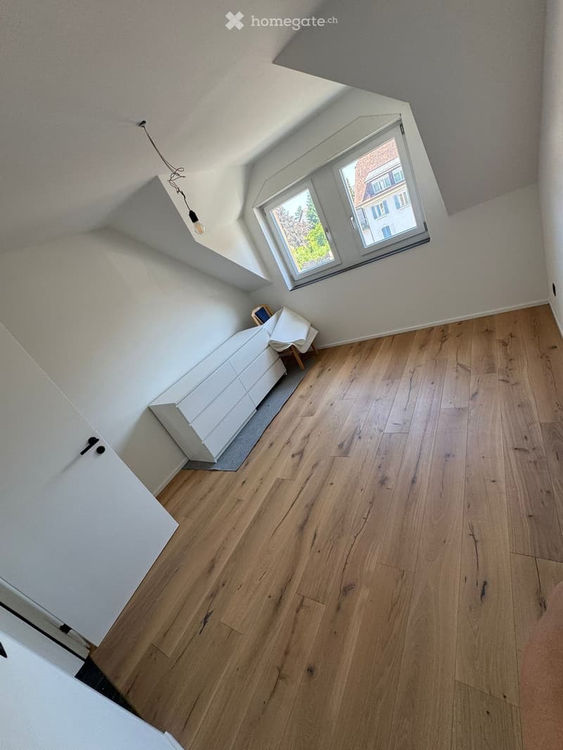 Bright, minimalist room with wooden flooring, white walls, and a large window offering a view of the outside. Features a white sofa and a small wooden stool.