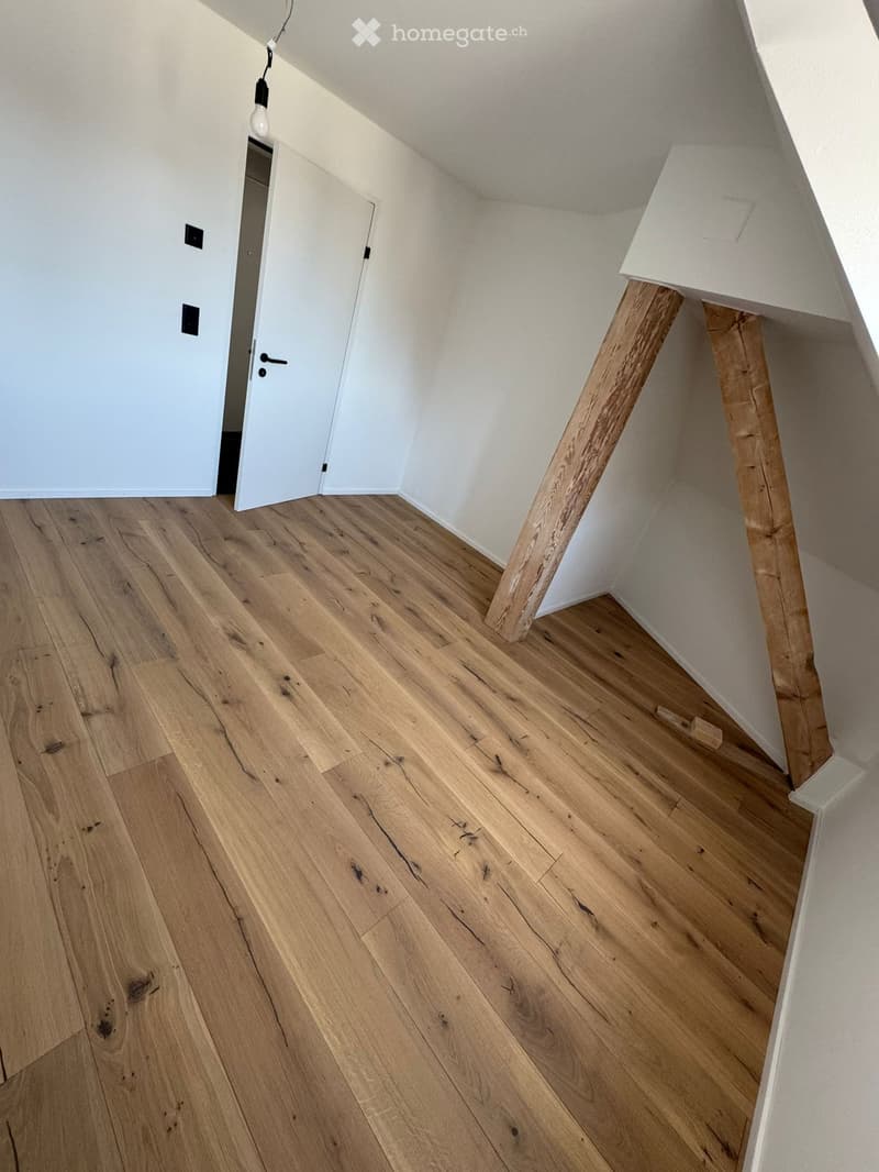 Empty room with wooden flooring and white walls, featuring a partially visible door and a hanging light fixture.