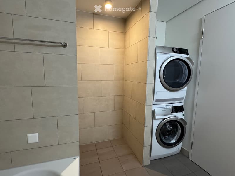 Modern laundry room with tiled walls, a washing machine, and a dryer, featuring a built-in shelf and a light fixture.