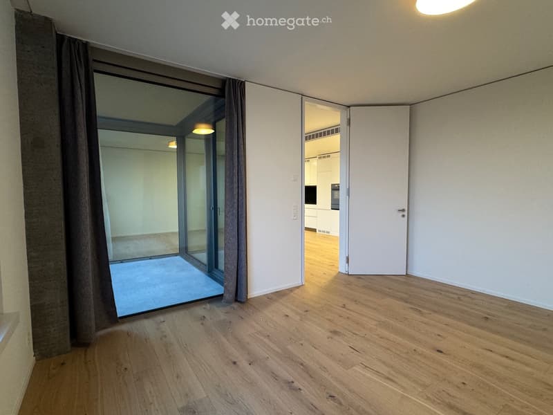 Empty room with wooden flooring, large glass sliding doors leading to a balcony, and an open doorway to another room.