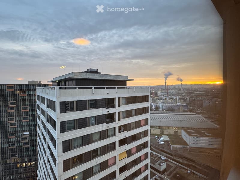 Modern high-rise building with multiple floors, large windows, and balconies overlooking a cityscape at sunset.